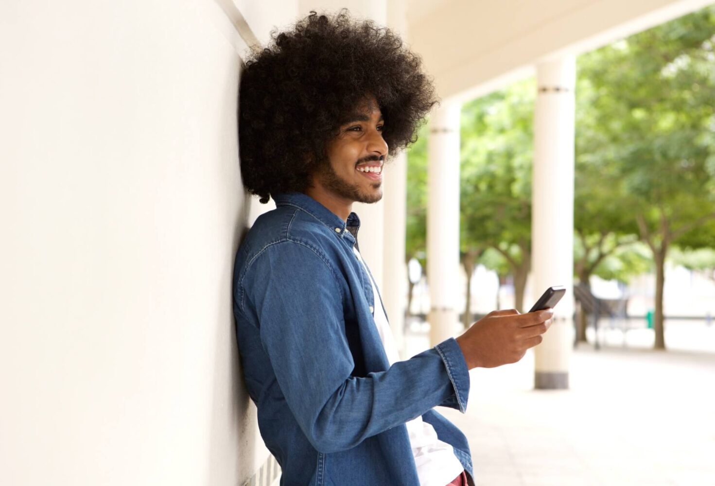 A young man with curly hair and a denim shirt smiles whilst using his mobile phone outdoors, highlighting mobile IT security support.